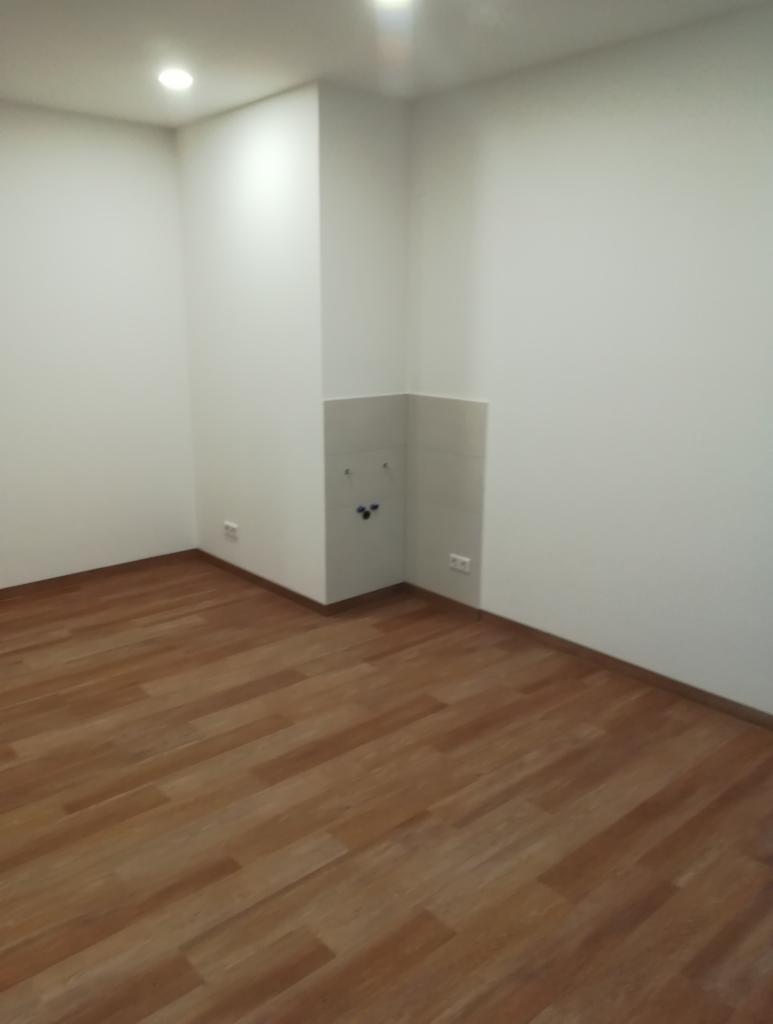 Puste, świeżo wykończone wnętrze z drewnianą podłogą, białymi ścianami i przygotowanym miejscem na instalację sanitarną.