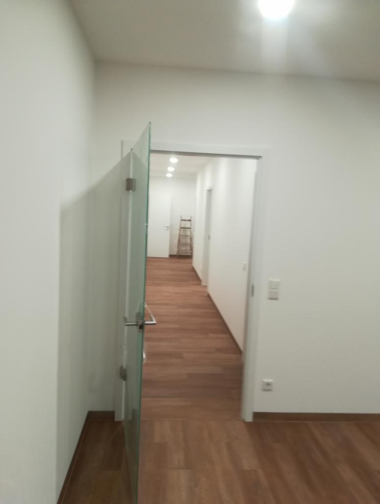 Wnętrze wyremontowanego mieszkania z drewnianą podłogą, widok na korytarz z otwartymi, szklanymi drzwiami i drabinką w tle, białe ściany i sufit z oświetleniem punktowym.