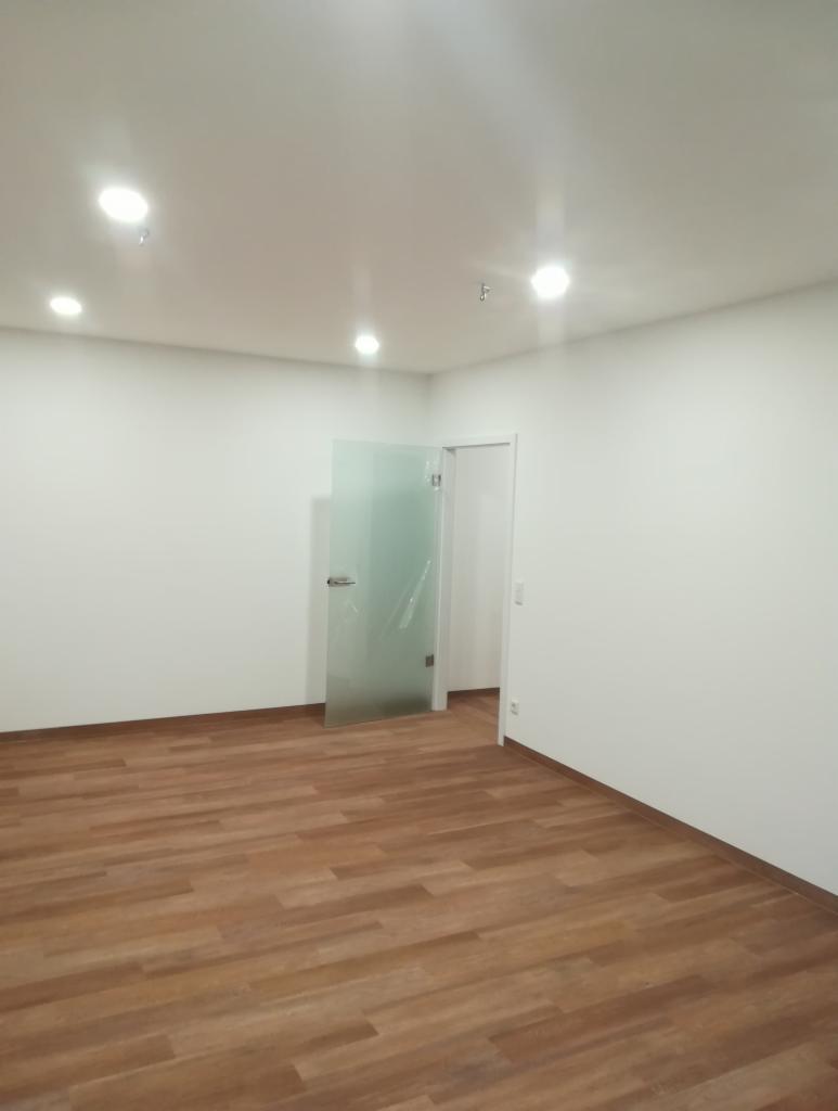 Puste, jasne pomieszczenie z drewnianą podłogą, białymi ścianami i szklanymi drzwiami, oświetlone punktowymi lampami sufitowymi. Folia ochronna wciąż na szklanych drzwiach.