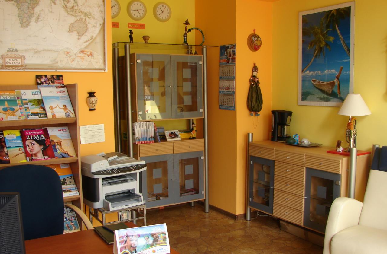 Wnętrze biura z mapą świata, zegarami wskazującymi różne strefy czasowe, regałem z folderami turystycznymi i meblami w odcieniach żółci i szarości.