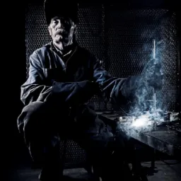 Pracownik w kombinezonie i masce spawalniczej, z wąsami, siedzi na krześle podczas spawania metalowej konstrukcji, widoczne iskry i dym, oświetlenie punktowe tworzy dramatyczny efekt.