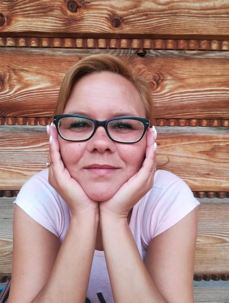 Uśmiechnięta kobieta w okularach opiera podbródek na dłoniach, na tle drewnianej ściany z widocznymi słojami.