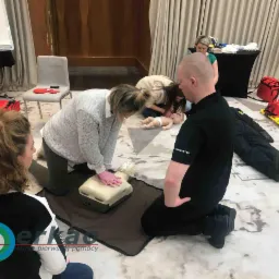 Grupa osób uczestniczy w symulacji resuscytacji krążeniowo-oddechowej na manekinie, pod okiem instruktora z napisem 'INSTRUKTOR' na koszulce, w sali konferencyjnej z tablicą flipchart i sprzętem...