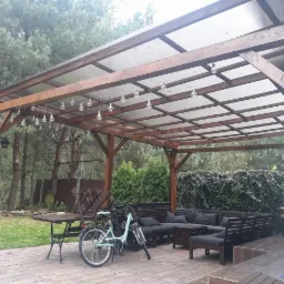 Zadaszona altana z drewnianą konstrukcją, oświetleniem w postaci wiszących żarówek, betonowym tarasem i meblami ogrodowymi, w tle drzewa i krzewy.