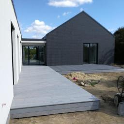 Mateusz Piechota Magiczny Taras. - Szary drewniany taras przy nowoczesnym domu z ciemnej cegły w trakcie budowy, widoczny fragment trawnika i narzędzia.