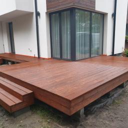 Mateusz Piechota Magiczny Taras. - Nowoczesny taras drewniany z brązowych desek, z trzema stopniami prowadzącymi do drzwi balkonowych, otoczony wilgotnym podłożem i zielenią.