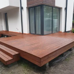 Nowoczesny taras drewniany z brązowych desek, z trzema stopniami prowadzącymi do drzwi balkonowych, otoczony wilgotnym podłożem i zielenią.