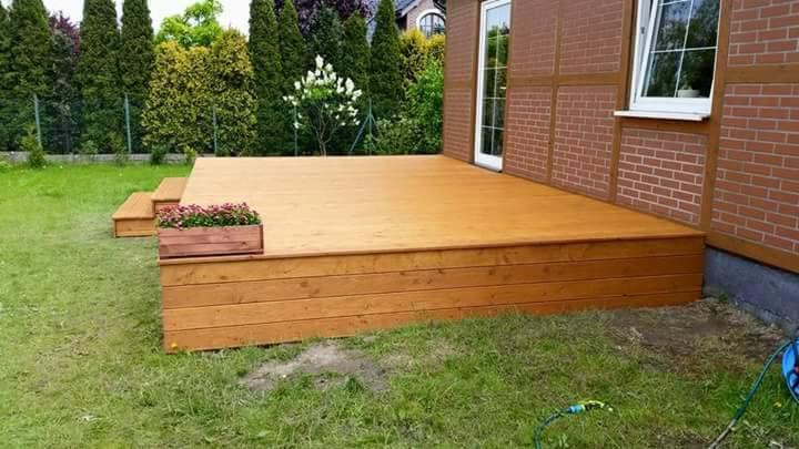 Nowo wybudowany, drewniany podest tarasowy z dwoma stopniami, skrzynką z kwiatami i widokiem na trawnik oraz elewację budynku z cegły.