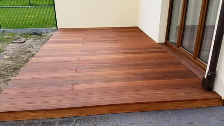 Wykończony taras z desek w odcieniach brązu przylegający do domu z drewnianą ramą okienną i brązową rynną.