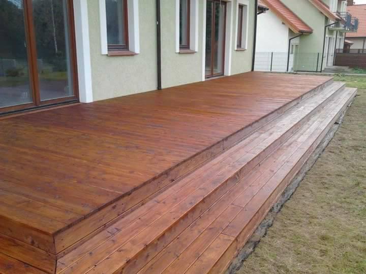 Nowoczesny, drewniany taras z trzema stopniami prowadzącymi do domu, wykończony w ciepłym, brązowym odcieniu, z widoczną strukturą drewna i gładką powierzchnią.