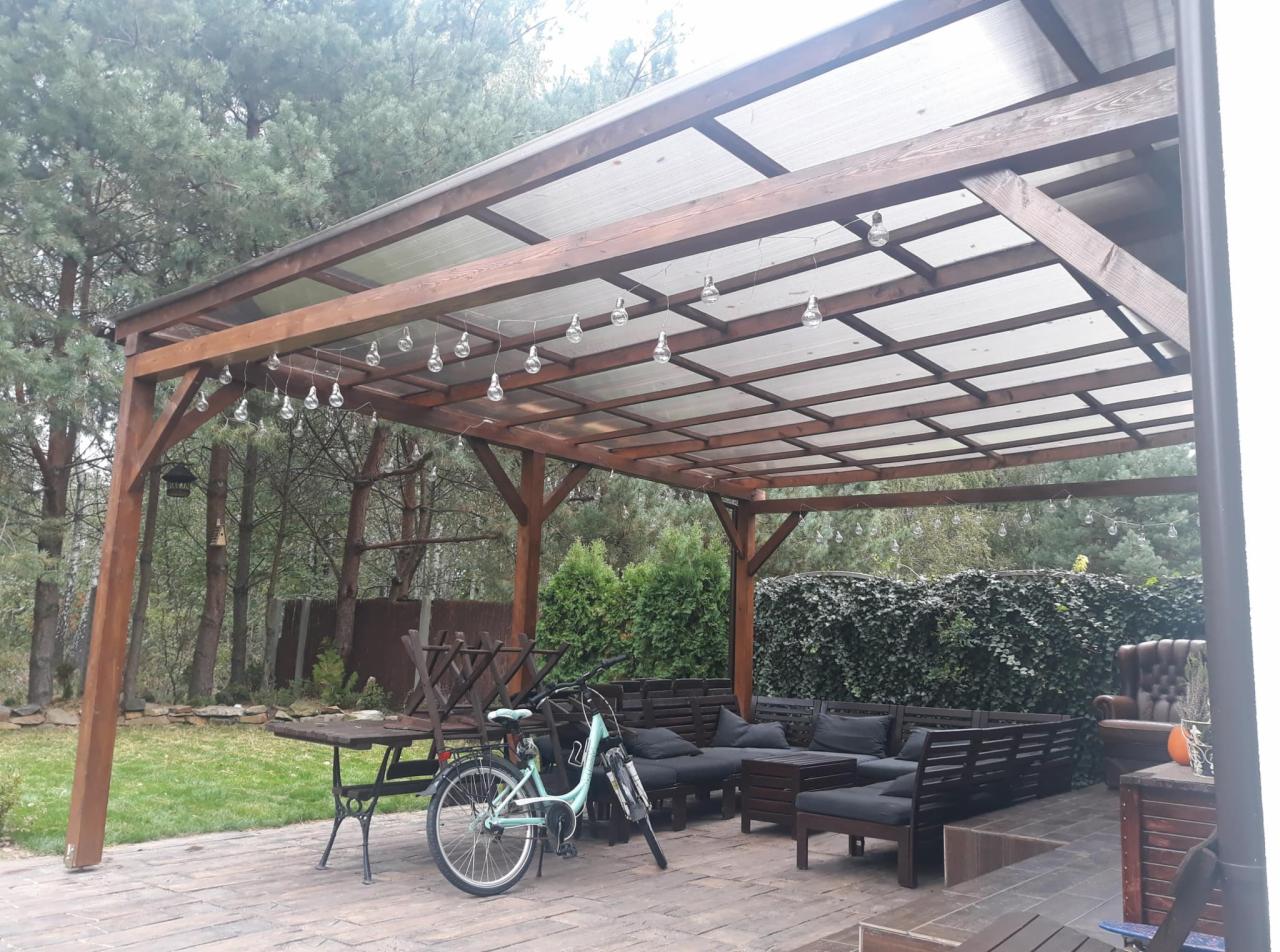 Zadaszona altana z drewnianą konstrukcją, oświetleniem w postaci wiszących żarówek, betonowym tarasem i meblami ogrodowymi, w tle drzewa i krzewy.