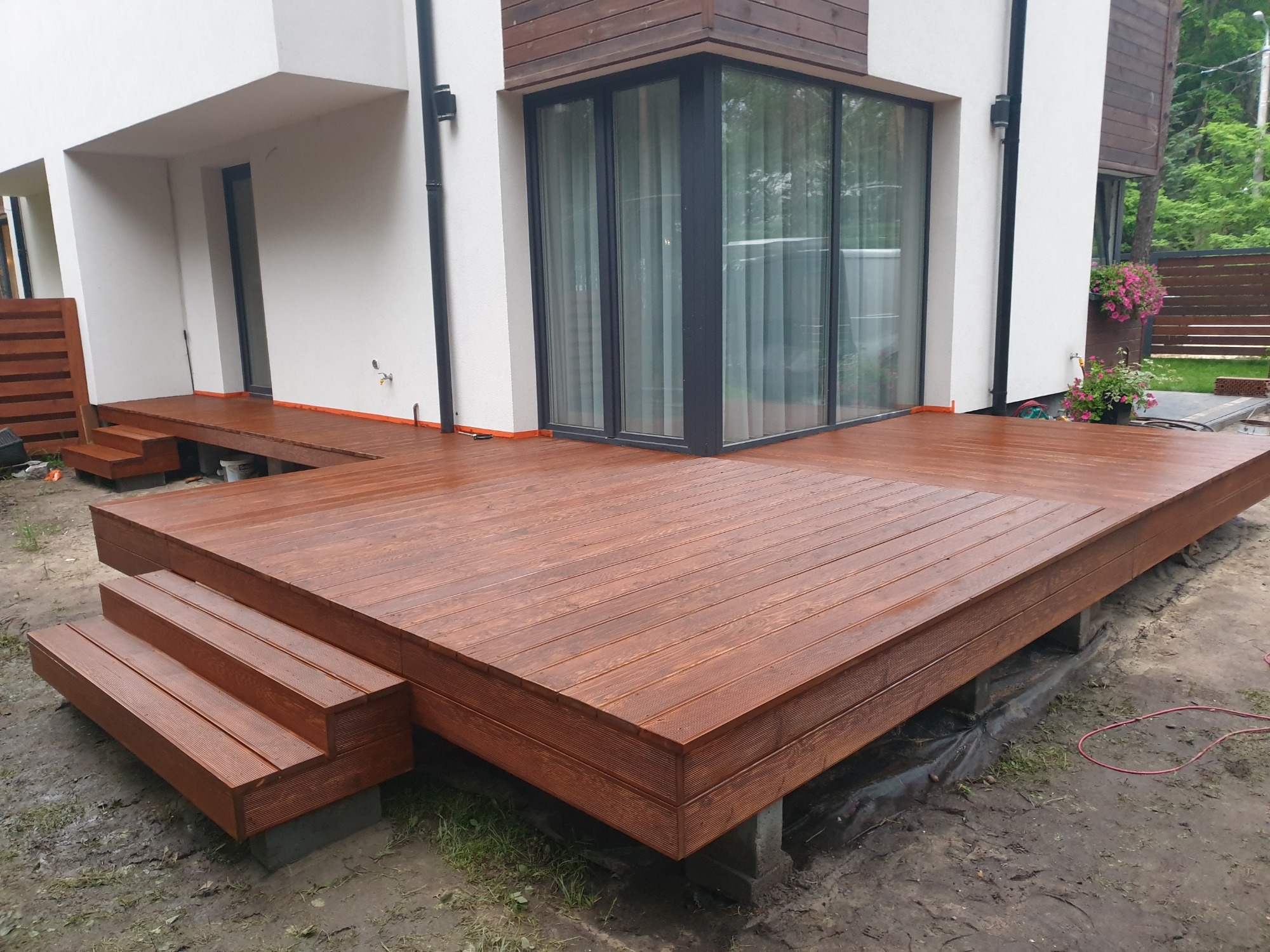 Nowoczesny taras drewniany z brązowych desek, z trzema stopniami prowadzącymi do drzwi balkonowych, otoczony wilgotnym podłożem i zielenią.