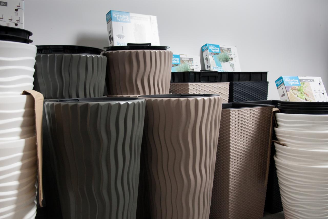 Różnokolorowe donice z tworzywa sztucznego o falistej i plecionej fakturze, ustawione obok siebie, z pudełkami systemów nawadniających w tle.