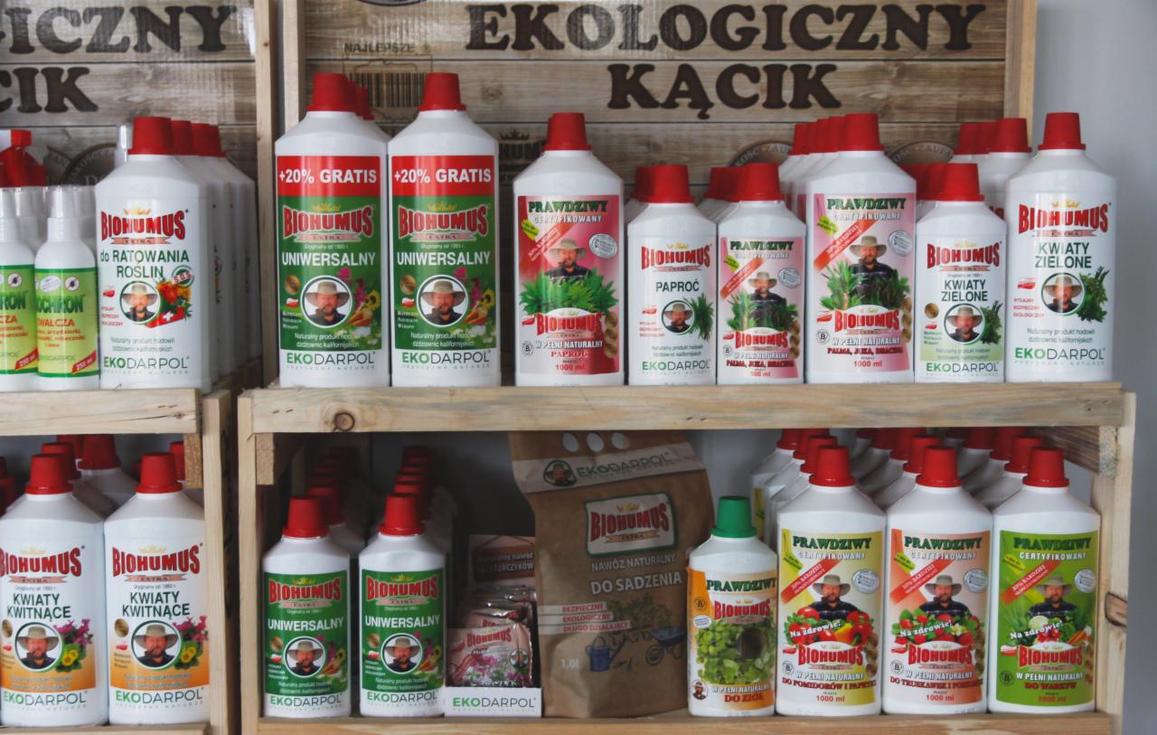 Półki sklepowe wypełnione różnymi rodzajami płynnych preparatów w białych butelkach z czerwonymi i zielonymi nakrętkami marki Ekodarpol, w tym Biohumus do różnych roślin, oraz papierowa torba...