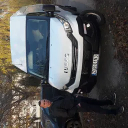 Biały bus marki Iveco z rejestracją ZGR 01N3, zaparkowany na poboczu, obok stoi mężczyzna w ciemnej kurtce.