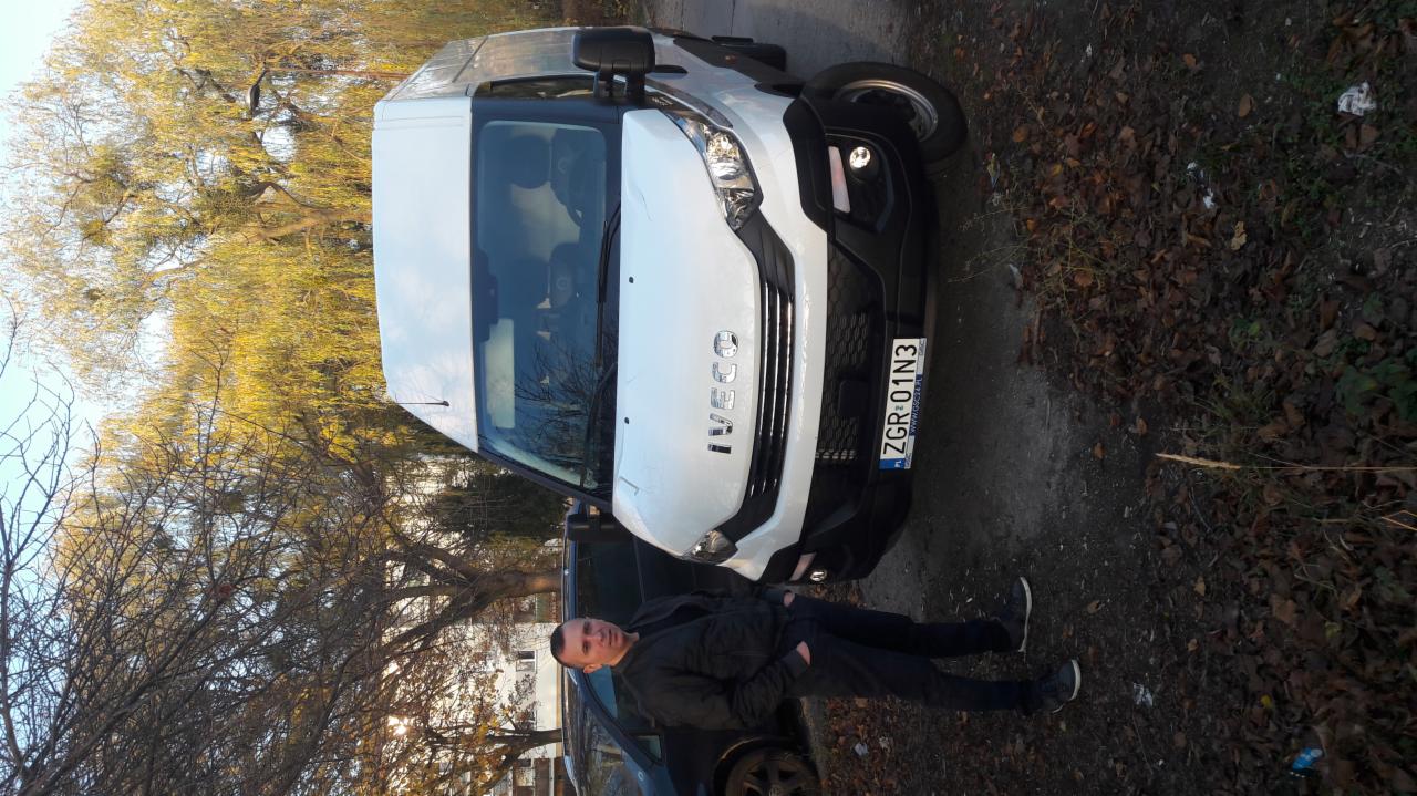 Biały bus marki Iveco z rejestracją ZGR 01N3, zaparkowany na poboczu, obok stoi mężczyzna w ciemnej kurtce.