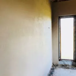 Świeżo nałożony tynk maszynowy na ścianie wewnętrznej, widoczne ślady po narzędziach, surowy stan wykończenia z oknem w tle, widoczne elementy konstrukcji stropu.
