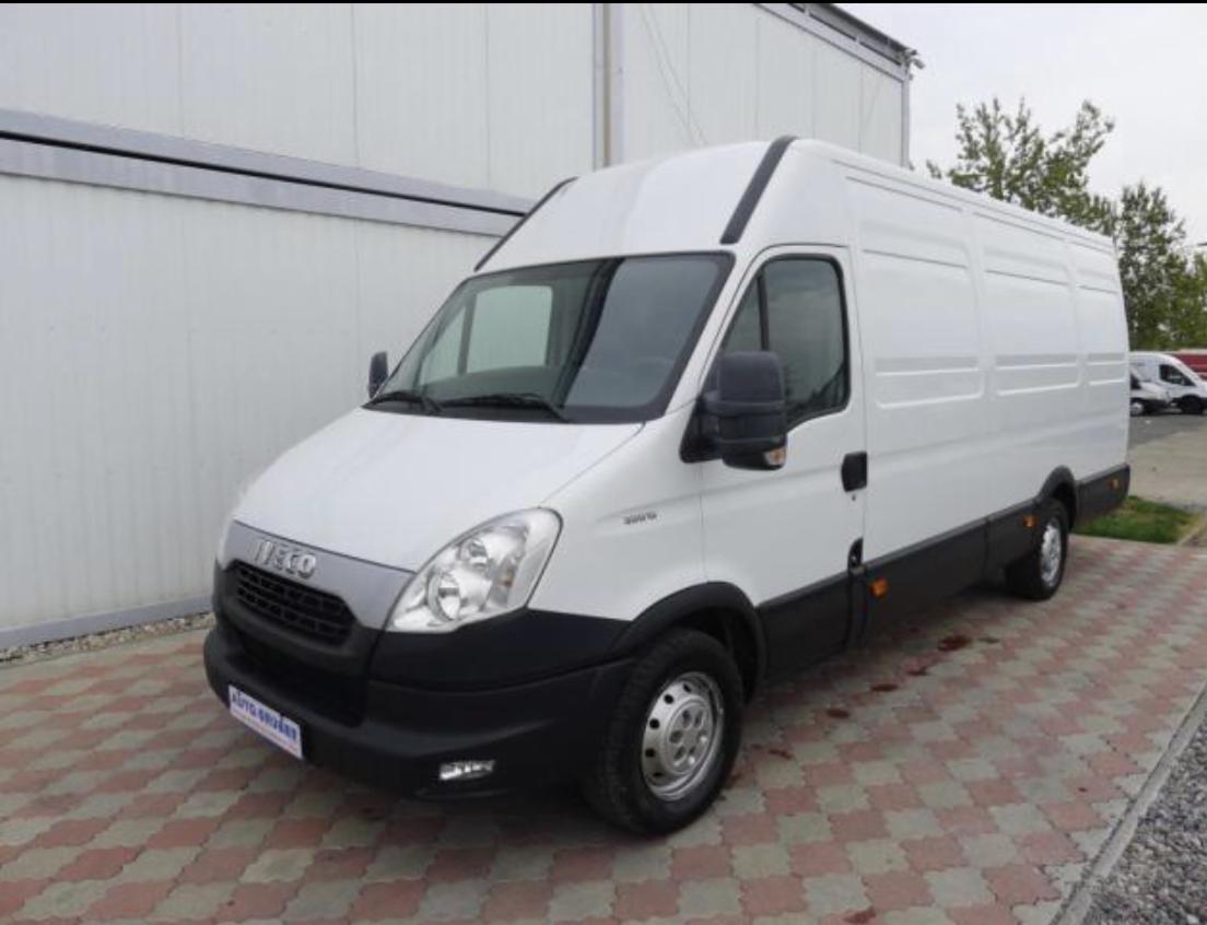 Biały samochód dostawczy marki Iveco z czarnym pasem u dołu, zaparkowany na kostce brukowej w pobliżu budynku magazynowego.