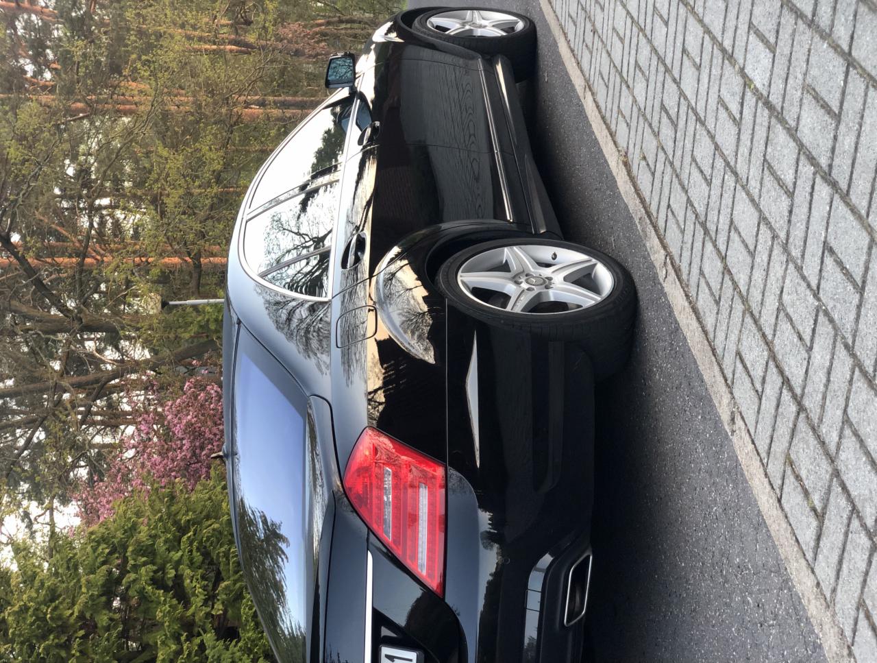 Czarny samochód osobowy marki Mercedes zaparkowany na brukowanej drodze, z widocznym fragmentem tylnej lampy, koła i refleksami w karoserii, na tle drzew i krzewów.