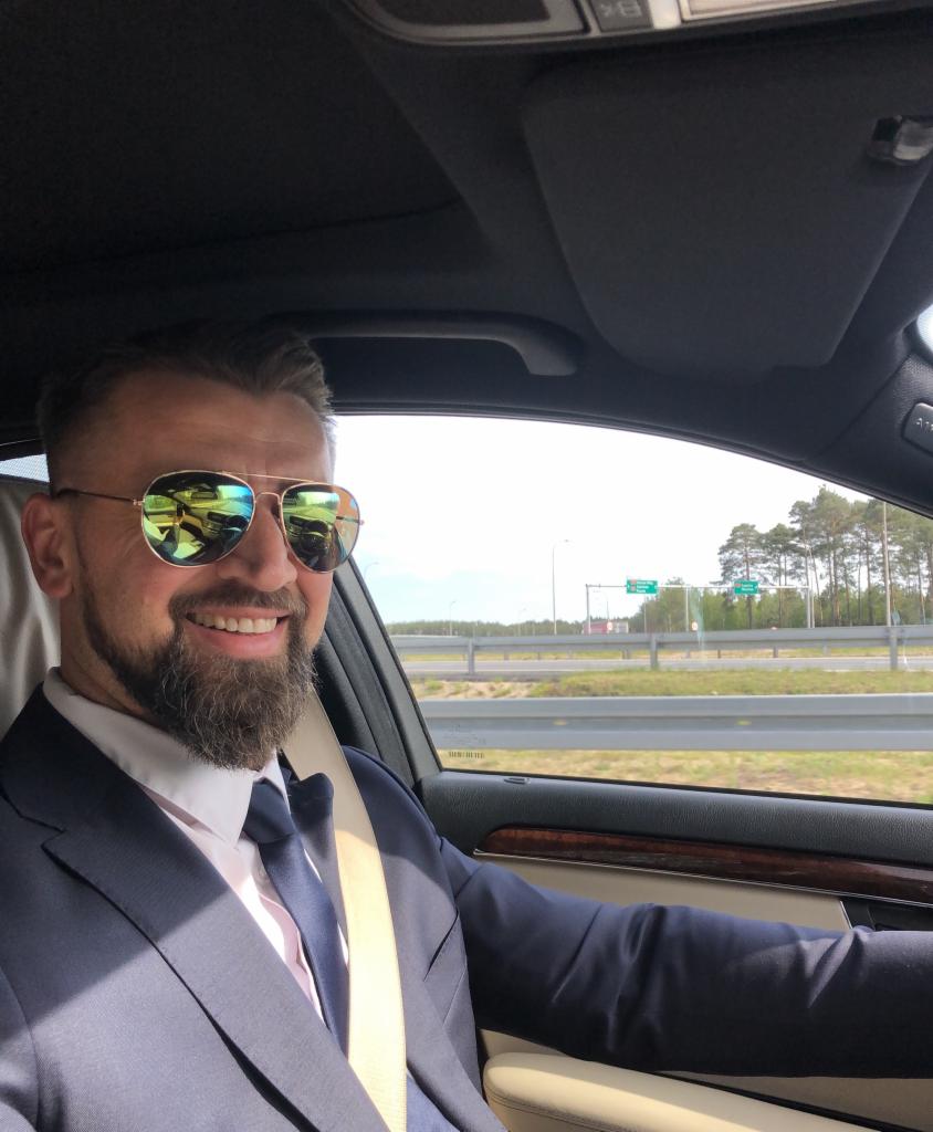 Uśmiechnięty mężczyzna w garniturze i okularach przeciwsłonecznych siedzi za kierownicą samochodu, widok na autostradę za oknem.