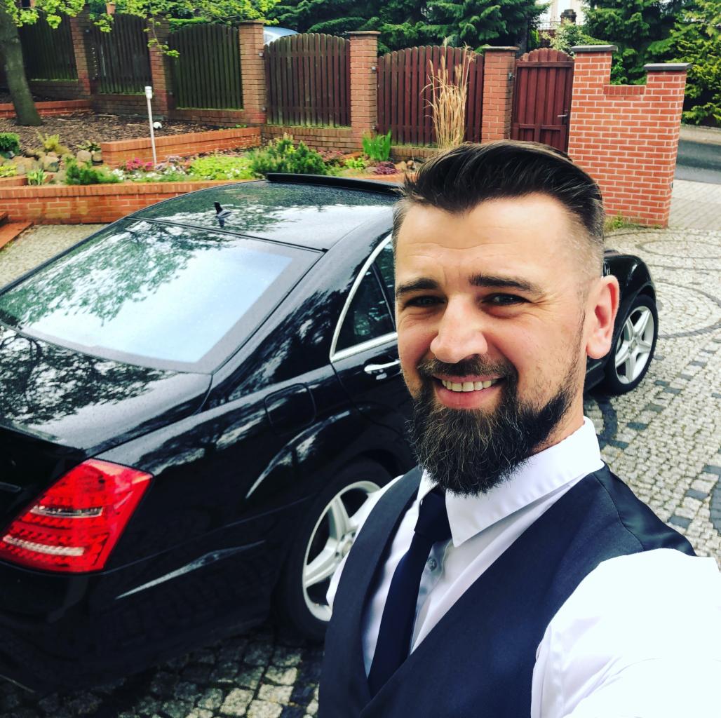 Uśmiechnięty mężczyzna w kamizelce i krawacie pozuje na tle czarnej limuzyny zaparkowanej na brukowanym podjeździe przed domem z ceglanym ogrodzeniem.