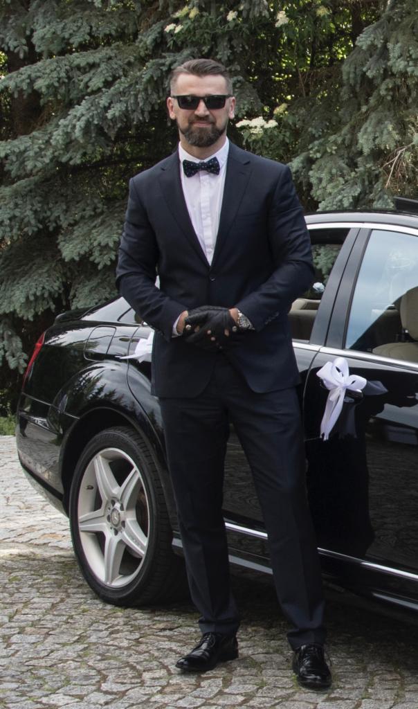 Elegancki mężczyzna w garniturze i muszce, w skórzanych rękawiczkach, stoi obok czarnego samochodu z ozdobną białą kokardą na klamce, prawdopodobnie przygotowanego do ślubu.