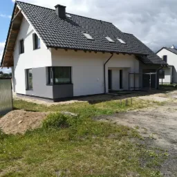 Nowoczesny dom jednorodzinny z szarą podmurówką i białymi ścianami, czarną dachówką i oknami w szarej ramie, widoczny w trakcie prac wykończeniowych na zewnątrz.