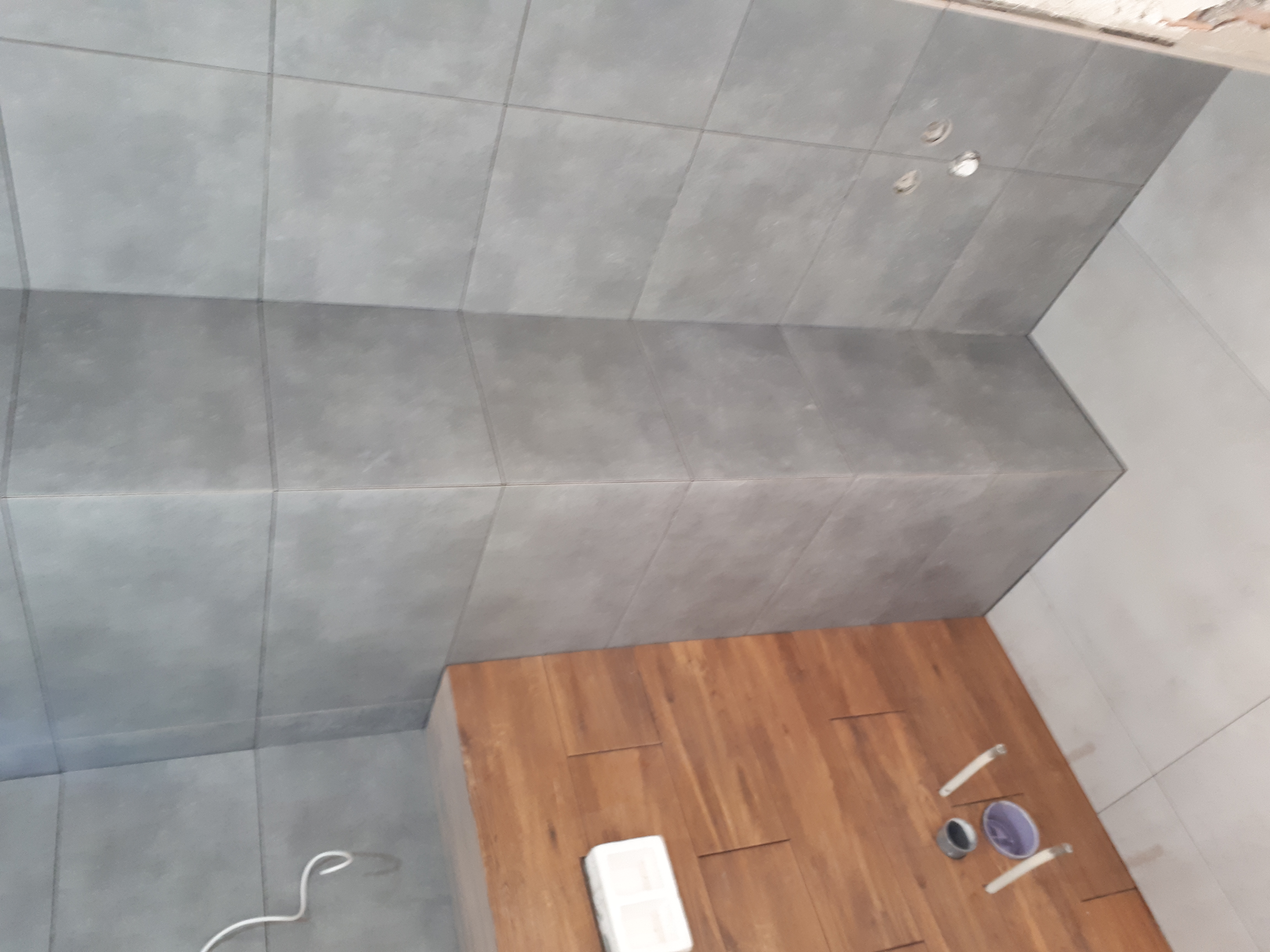 Szare płytki ścienne ułożone do połowy wysokości pomieszczenia, kontrastujące z drewnopodobną podłogą. Widoczne przygotowane materiały i narzędzia glazurnicze.