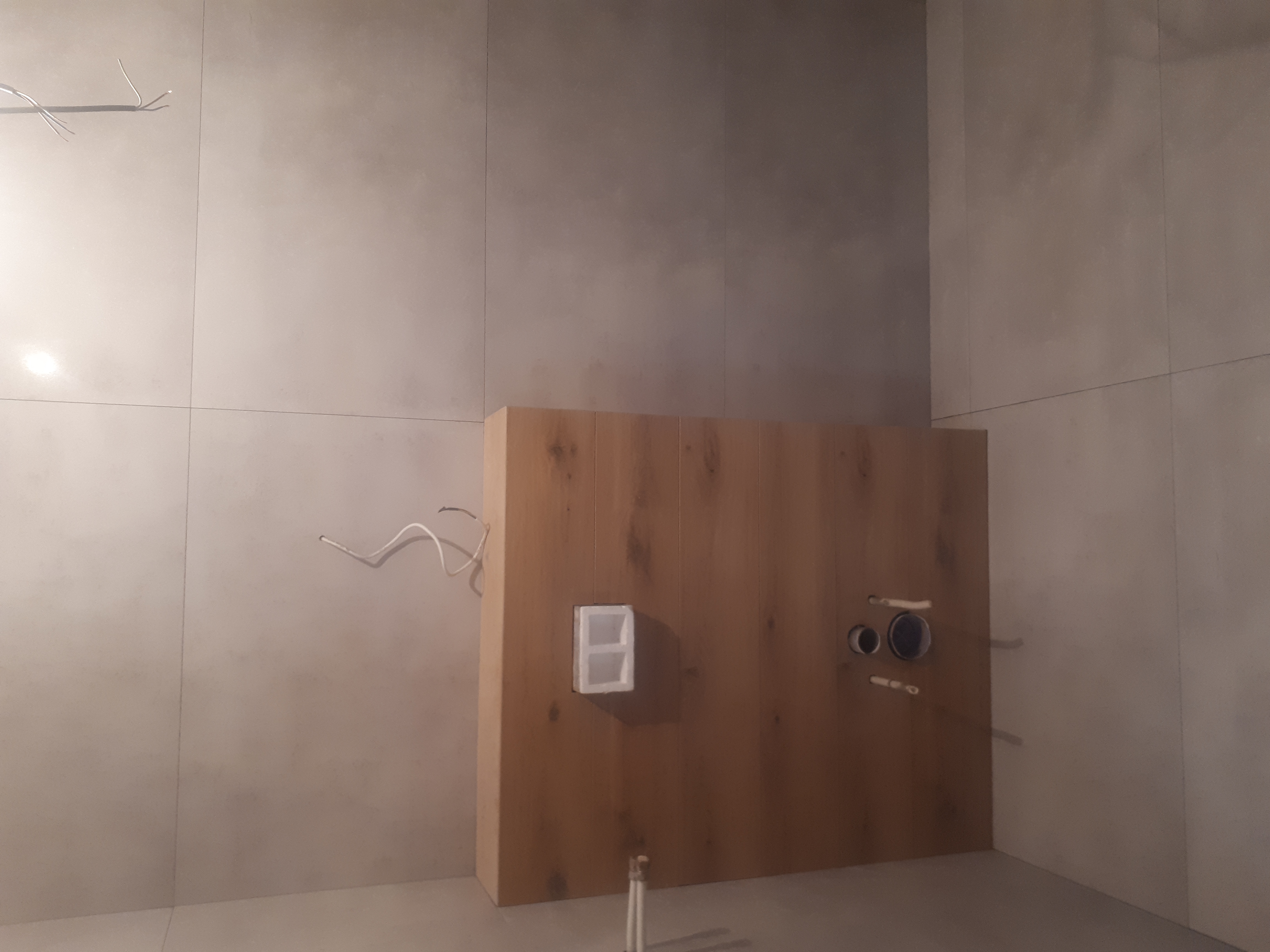 Ściana łazienki z szarymi płytkami, drewnianą zabudową stelaża WC z widocznymi przyłączami hydraulicznymi i elektrycznymi, w trakcie montażu.