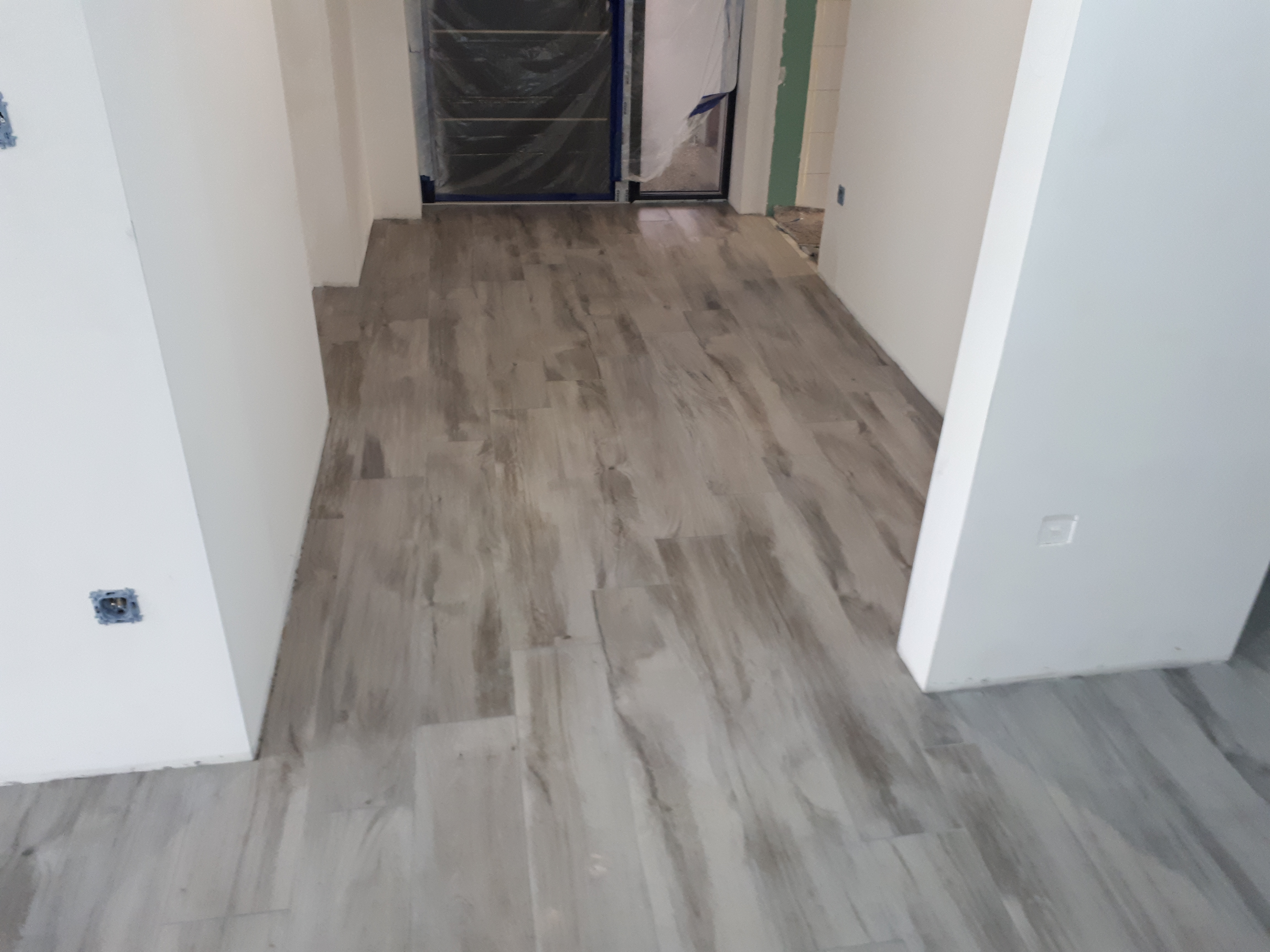 Ułożona podłoga z płytek imitujących drewno w korytarzu, z widocznymi puszkami elektrycznymi na ścianie i zabezpieczonymi folią drzwiami w tle.