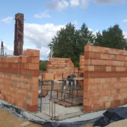 Wznoszone ściany domu z czerwonej cegły ceramicznej na fundamencie, widoczne rusztowania, komin z cegły obrośnięty bluszczem oraz pracownik w tle.