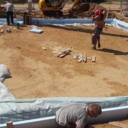 NewHome Łukasz Szkulimowski - Przygotowanie szalunku i zbrojenia pod fundament domu jednorodzinnego, widoczny teren budowy, koparka i pracownicy w trakcie pracy.