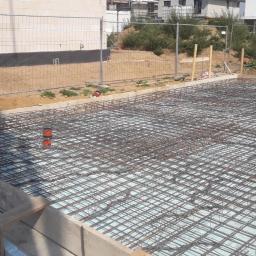 NewHome Łukasz Szkulimowski - Zbrojenie fundamentu na warstwie izolacji termicznej, widoczne szalunki i wystające rury instalacyjne, w tle ogrodzenie placu budowy i budynki w trakcie budowy.