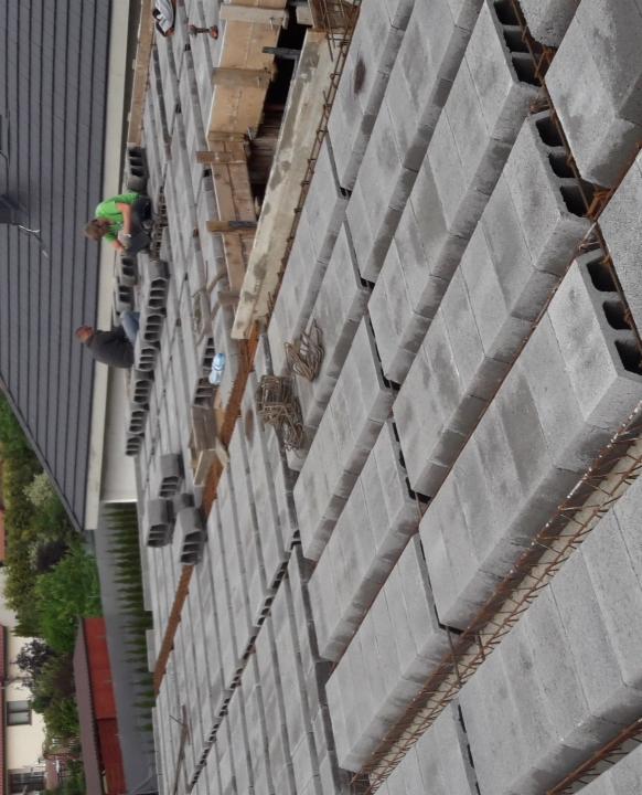 Budowa ściany z szarych bloczków betonowych, widoczna stalowa siatka zbrojeniowa, dwóch pracowników w trakcie pracy, widok z góry.