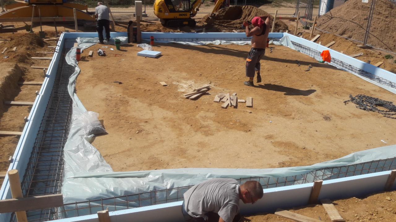 Przygotowanie szalunku i zbrojenia pod fundament domu jednorodzinnego, widoczny teren budowy, koparka i pracownicy w trakcie pracy.