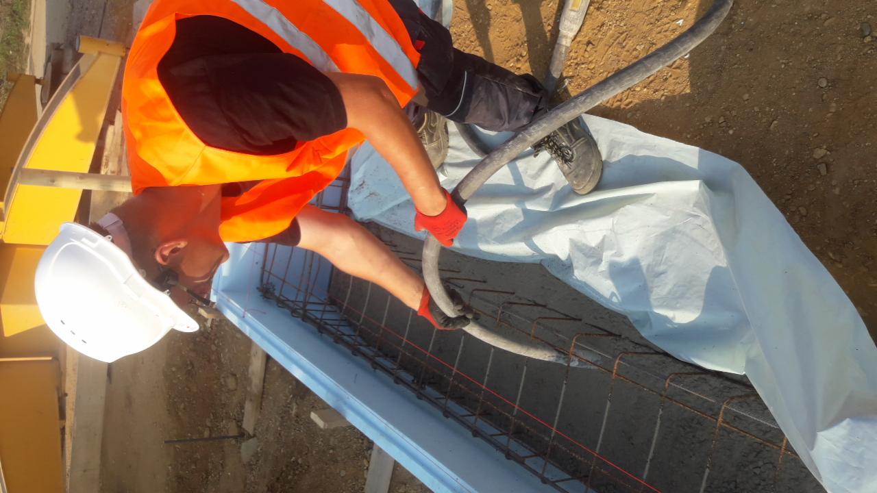 Pracownik w kasku i kamizelce odblaskowej wylewa beton z węża do szalunku zbrojeniowego na budowie.