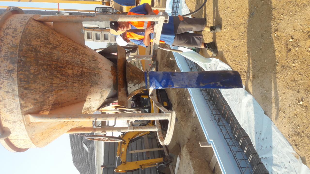 Pracownik w kamizelce odblaskowej nadzoruje wylewanie betonu z brudnej gruszki betoniarki do szalunku zbrojeniowego na fundamencie, widoczna koparka i fragment budynku w tle.