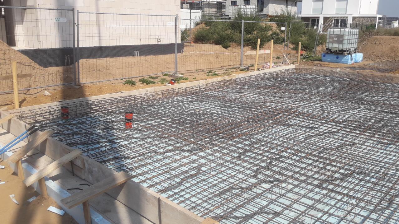 Zbrojenie fundamentu na warstwie izolacji termicznej, widoczne szalunki i wystające rury instalacyjne, w tle ogrodzenie placu budowy i budynki w trakcie budowy.