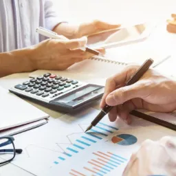 Analiza wykresu z danymi finansowymi przy użyciu kalkulatora i tabletu, dwie osoby, ołówek w dłoni, ciepłe światło.