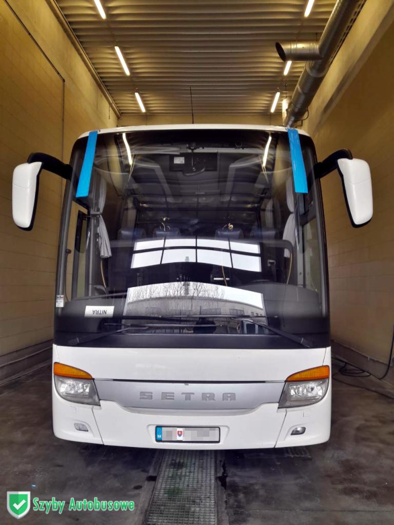 Przód białego autobusu marki Setra z niebieską taśmą na szybie, stojącego w hali z widocznymi instalacjami na suficie.