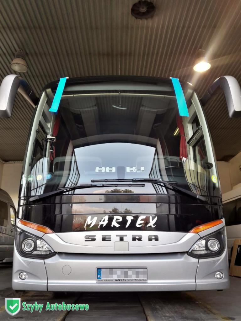Przód srebrnego autobusu Setra Martex z niebieską taśmą na szybie czołowej, stojącego w warsztacie z widocznymi lampami pod sufitem.