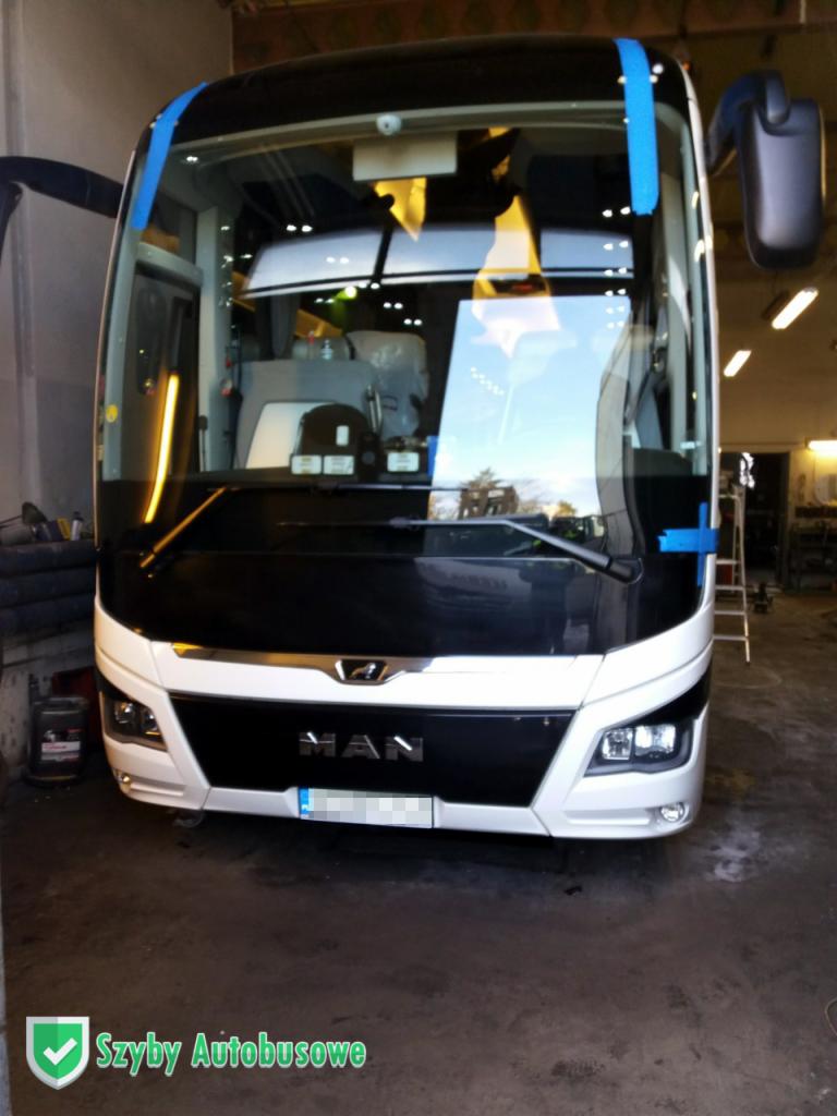 Przód autobusu MAN z nową szybą czołową, zabezpieczoną niebieską taśmą, w warsztacie.