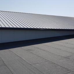 MiKAR - Widok z góry na dach hali z blachy trapezowej, z widoczną ścianą z płyt warstwowych i pracownikiem na dachu, stojącym na podwyższeniu.