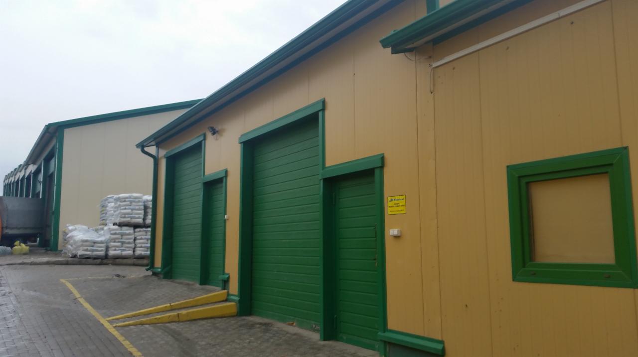 Zewnętrzny widok hali przemysłowej z żółtymi ścianami z płyty warstwowej i zielonymi bramami rolowanymi, widoczne worki z materiałem budowlanym i rampy załadunkowe.