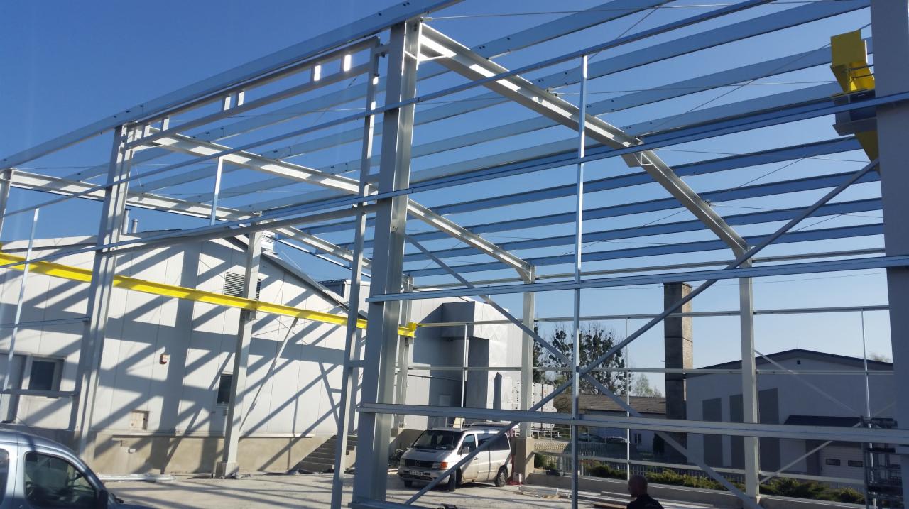 Konstrukcja stalowa hali w trakcie montażu, widoczne elementy nośne, słupy i belki, na tle budynek w trakcie budowy i zaparkowany samochód dostawczy, ujęcie z dołu.