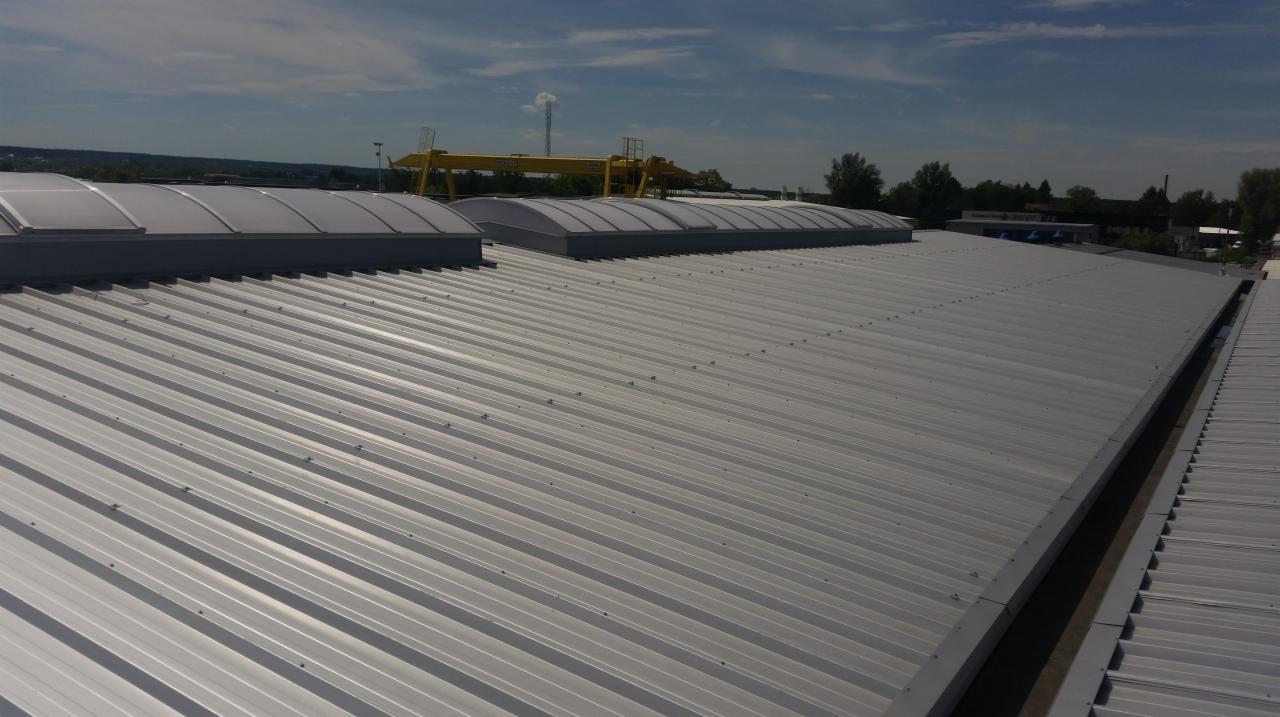 Długi, srebrny dach z blachy trapezowej z kilkoma świetlikami kopułowymi, widoczny suwnica bramowa w tle.