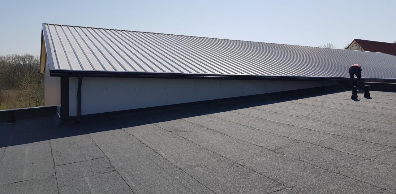 Widok z góry na dach hali z blachy trapezowej, z widoczną ścianą z płyt warstwowych i pracownikiem na dachu, stojącym na podwyższeniu.