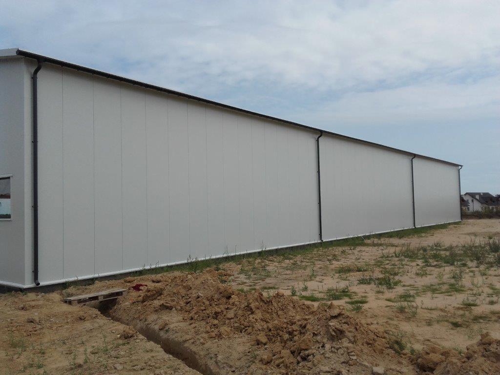 Długi budynek przemysłowy z białych płyt warstwowych, widoczny z zewnątrz, z ziemią i skąpą roślinnością na pierwszym planie, pochmurne niebo w tle.