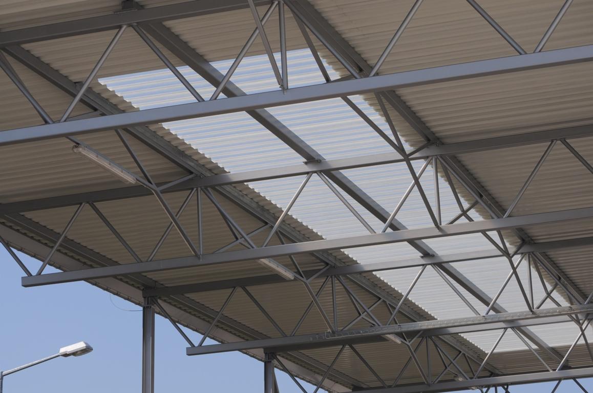 Widok z dołu na konstrukcję stalową z zamontowanymi płytami warstwowymi przepuszczającymi światło, oświetlenie ledowe i fragment słupa oświetleniowego na tle błękitnego nieba.