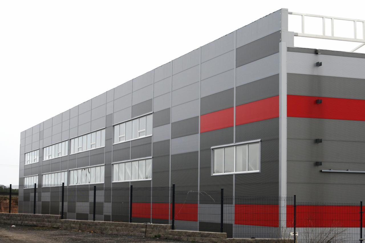 Nowoczesna hala przemysłowa z szarymi i czerwonymi płytami warstwowymi, widok z zewnątrz, ogrodzona siatką.